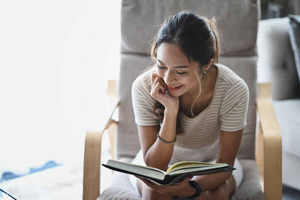Mujer joven satisfecha leyendo libro en sillón mientras pasa tiempo libre en casa. - Foto, imagen