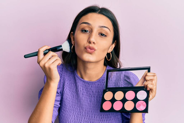 Young hispanic woman holding makeup brush and blush looking at the camera blowing a kiss being lovely and sexy. love expression.  - Photo, Image