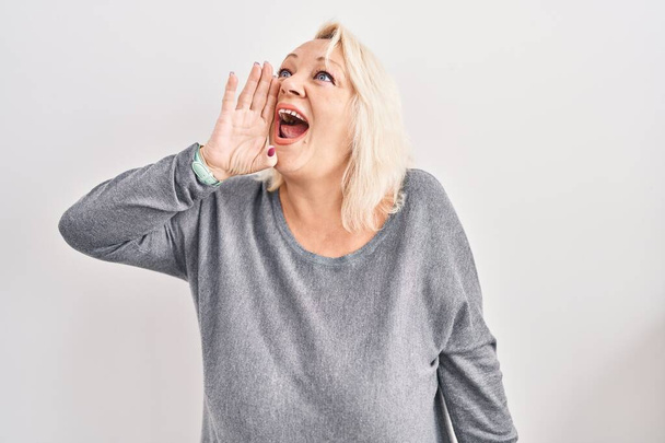 Middle age caucasian woman standing over white background shouting and screaming loud to side with hand on mouth. communication concept.  - Photo, Image
