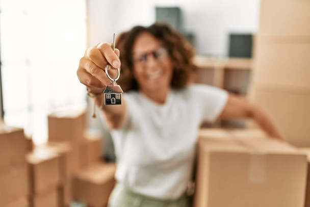 Middle age hispanic woman holding cardboard box and key of house at new home - Foto, Imagen