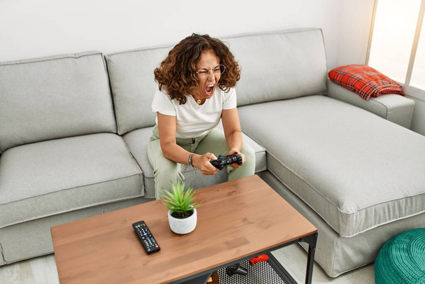 Mature hispanic woman screaming playing video games sitting on the sofa at home - Photo, Image