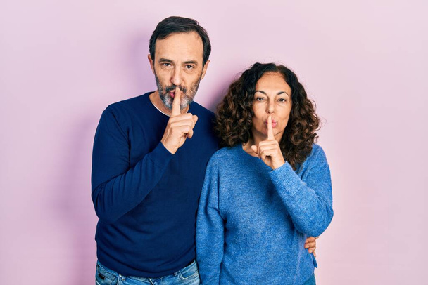 Middle age couple of hispanic woman and man hugging and standing together asking to be quiet with finger on lips. silence and secret concept.  - Photo, Image