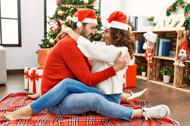 Middle age hispanic couple wearing christmas hat sitting on the floor and hugging at home. - Photo, Image