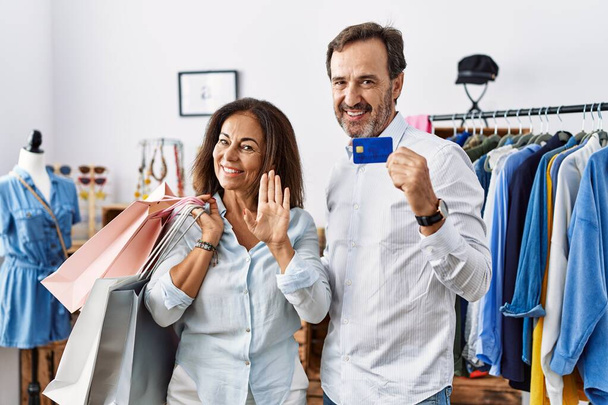 Ισπανόφωνο ζευγάρι μέσης ηλικίας κρατώντας τσάντες για ψώνια και πιστωτική κάρτα παραίτηση λέγοντας γεια χαρούμενος και χαμογελαστός, φιλική χειρονομία καλωσορίσματος  - Φωτογραφία, εικόνα