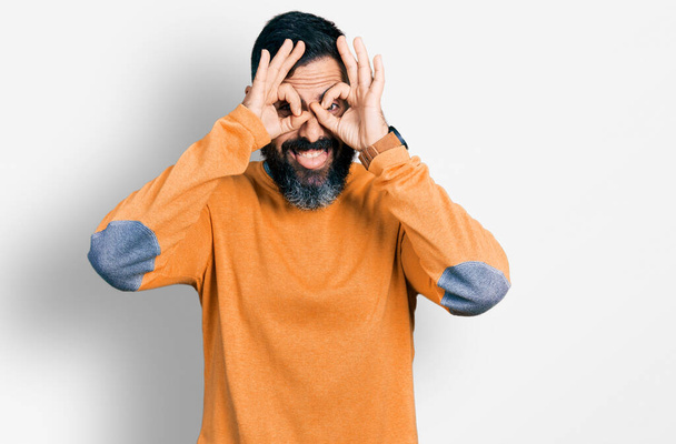 Hombre hispano con barba usando suéter casual de invierno haciendo buen gesto como binoculares sacando la lengua, ojos mirando a través de los dedos. expresión loca.  - Foto, imagen