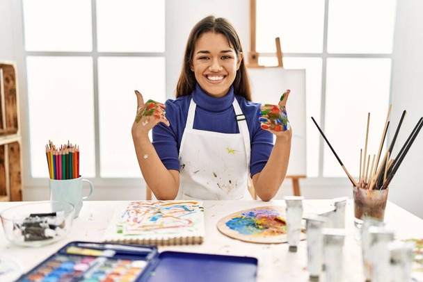 Νεαρή μελαχρινή γυναίκα στο στούντιο τέχνης με ζωγραφισμένα χέρια σημάδι επιτυχίας κάνει θετική χειρονομία με το χέρι, τους αντίχειρες επάνω χαμογελώντας και χαρούμενος. Χαρούμενη έκφραση και χειρονομία νικητή.  - Φωτογραφία, εικόνα