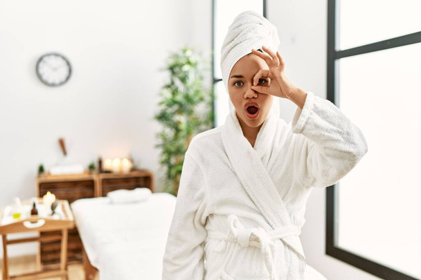 Young brunette woman wearing towel and bathrobe standing at beauty center doing ok gesture shocked with surprised face, eye looking through fingers. unbelieving expression.  - Photo, Image