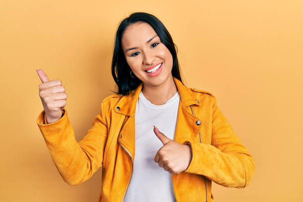 Mooie latino vrouw met neus piercing dragen gele leren jas wijzend naar de achterkant met de hand en duimen omhoog, glimlachend vol vertrouwen  - Foto, afbeelding