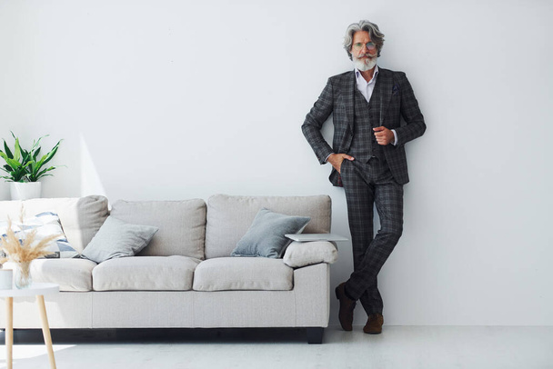 In elegant clothes. Standing against white wall. Senior stylish modern man with grey hair and beard indoors. - Foto, imagen