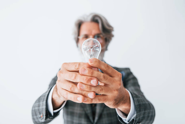 With light bulb in hands. Senior stylish modern man with grey hair and beard indoors. - Foto, Imagen
