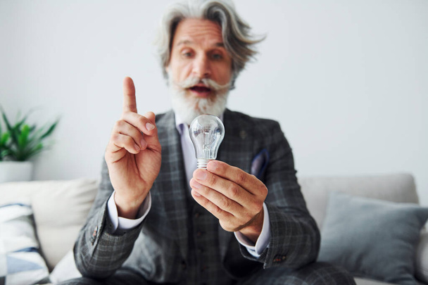 Sits on comfortable sofa in formal clothes. Senior stylish modern man with grey hair and beard indoors. - Photo, image