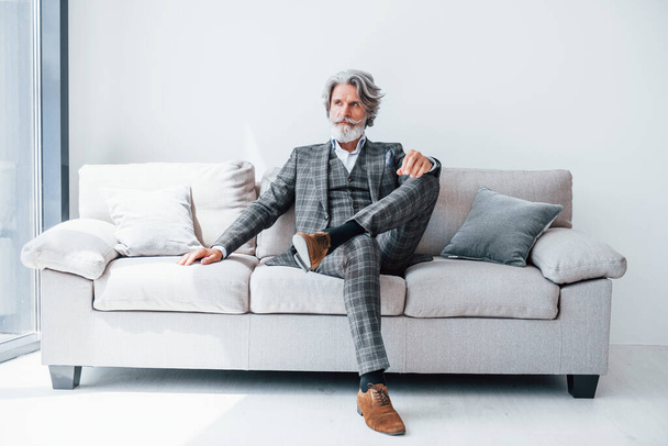 Sits on comfortable sofa in formal clothes. Senior stylish modern man with grey hair and beard indoors. - Фото, зображення