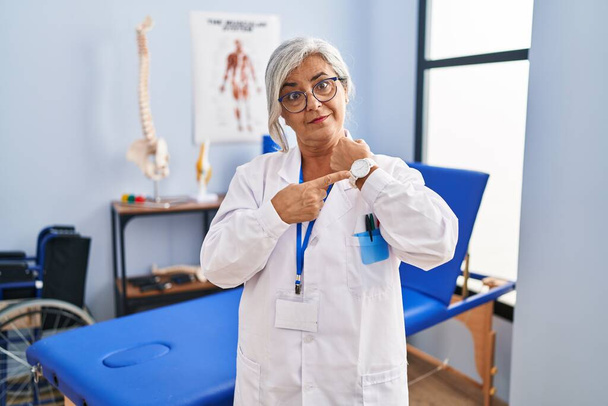 Middle age woman with grey hair working at pain recovery clinic in hurry pointing to watch time, impatience, looking at the camera with relaxed expression  - 写真・画像