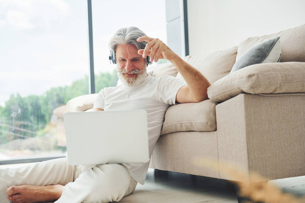 Sits on the ground near sofa with laptop and listens music. Senior stylish modern man with grey hair and beard indoors. - Φωτογραφία, εικόνα