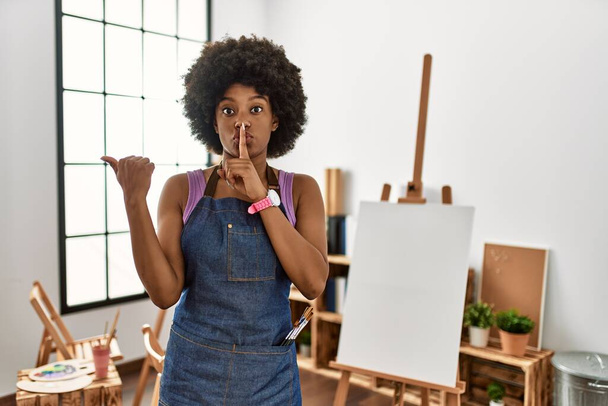 Fiatal afro-amerikai nő afro hajjal a művészeti stúdióban, aki azt kéri, hogy maradjon csendben, miközben az ujjai a kézfejével oldalra mutogatnak. csend és titkos koncepció.  - Fotó, kép
