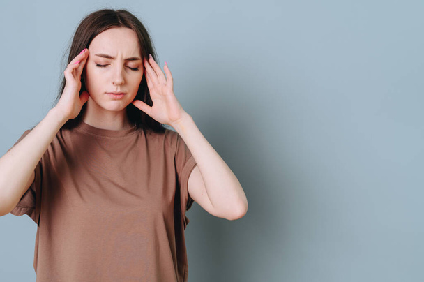 Portrait of a young girl in a brown T-shirt on a blue background, hands near the temples, closed eyes with a serious expression. The concept of escape from reality, a cure for headaches. - Photo, Image