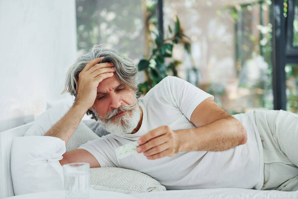 Sick senior stylish modern man with grey hair and beard indoors lying on the bed and feels bad. - Foto, Imagen