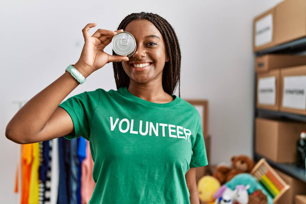 Mladý africký Američan žena na sobě dobrovolník uniforma držení konzervy jídlo přes oko v charitativním centru - Fotografie, Obrázek