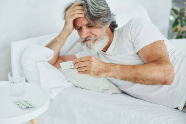 Sick senior stylish modern man with grey hair and beard indoors lying on the bed and feels bad. - Foto, imagen