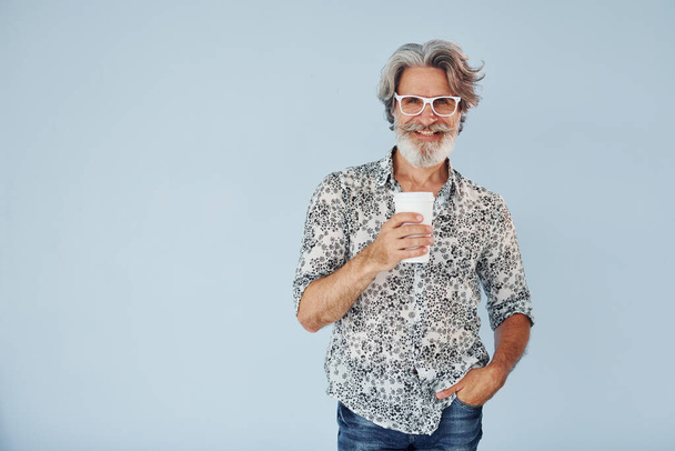 Чашка выпивки в руках. Старший стильный современный мужчина с седыми волосами и бородой в помещении. - Фото, изображение