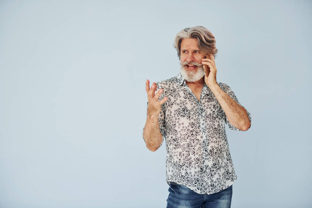 Having call when standing against wall. Senior stylish modern man with grey hair and beard indoors. - Фото, изображение