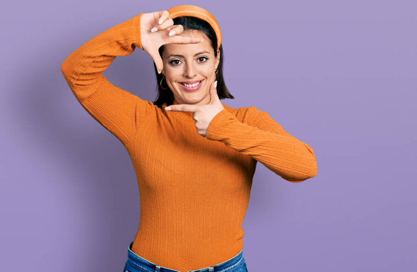 Ein junges hispanisches Mädchen in einem lässigen weißen T-Shirt, das lächelnd mit Händen und Fingern ein fröhliches Gesicht macht. Kreativität und Fotografiekonzept.  - Foto, Bild