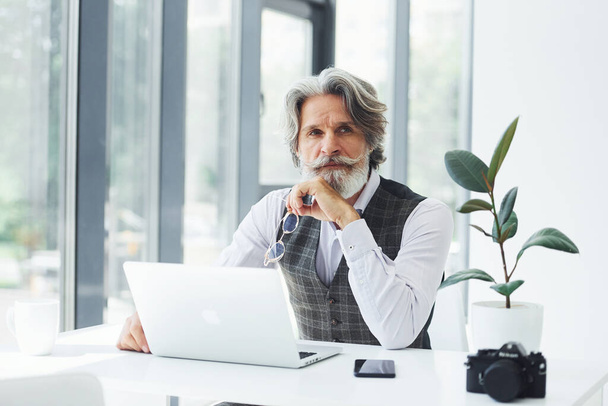 Businessman sits by table with laptop. Senior stylish modern man with grey hair and beard indoors. - 写真・画像