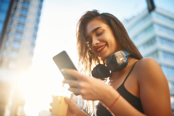 Atrakcyjna dziewczyna pijąca zimną kawę i patrząca na smartfona. Stylowy nastolatek z blizną rozmawiający z przyjaciółmi i uśmiechnięty. Czas letni w wielkim mieście. Dziewczyna na zachodzie słońca - Zdjęcie, obraz