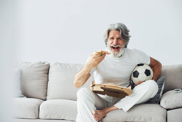 Soccer fan with pizza watches match. Senior stylish modern man with grey hair and beard indoors. - Zdjęcie, obraz