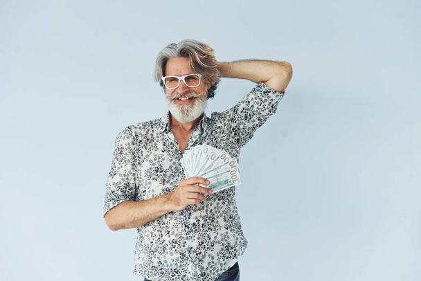 Senior stylish modern man with grey hair and beard indoors and holds cash. - Фото, изображение