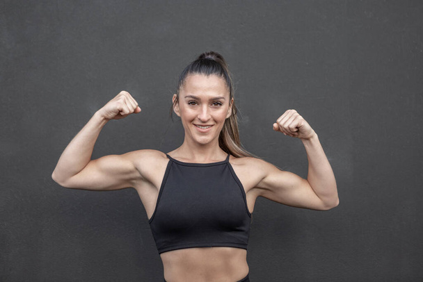 Atleta femenina musculosa positiva en top negro mirando a la cámara con sonrisa y mostrando bíceps mientras está de pie contra la pared negra durante el entrenamiento en el gimnasio - Foto, Imagen