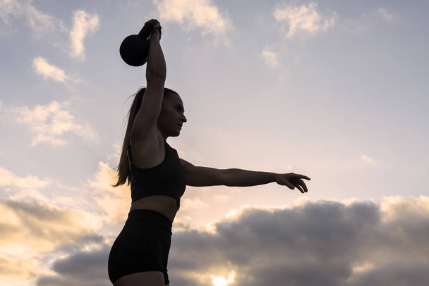 Niski kąt dopasowania kobiety sportowca w odzieży sportowej z końskim ogonem podnoszącym ramię z ciężkim dzwonkiem nad głową podczas ćwiczeń przeciwko zachmurzonemu niebu zachodu słońca - Zdjęcie, obraz