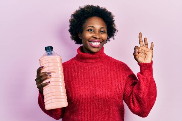 Genç Afro-Amerikalı kadın elinde deterjan şişesi tutuyor. Parmaklarıyla imza atıyor, gülümseyerek mükemmel bir sembol oluşturuyor.  - Fotoğraf, Görsel