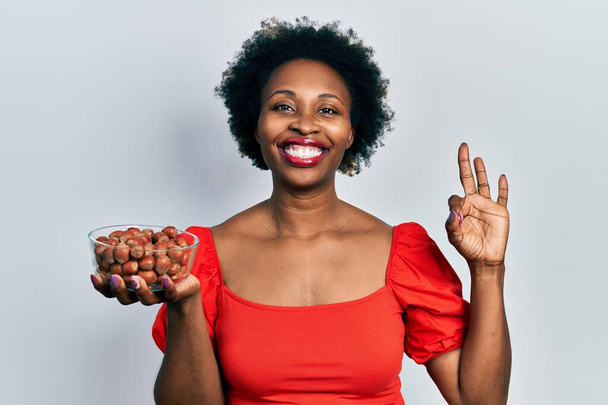 Jonge Afro-Amerikaanse vrouw met rauwe hazelnoten doen ok teken met de vingers, glimlachend vriendelijk gebaar uitstekende symbool  - Foto, afbeelding