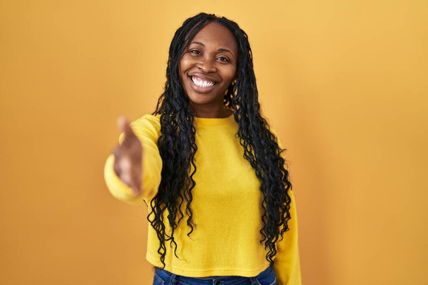 Αφρικανική γυναίκα στέκεται πάνω από κίτρινο φόντο χαμογελώντας φιλικό προσφέροντας χειραψία ως χαιρετισμό και φιλόξενο. επιτυχημένη επιχείρηση.  - Φωτογραφία, εικόνα