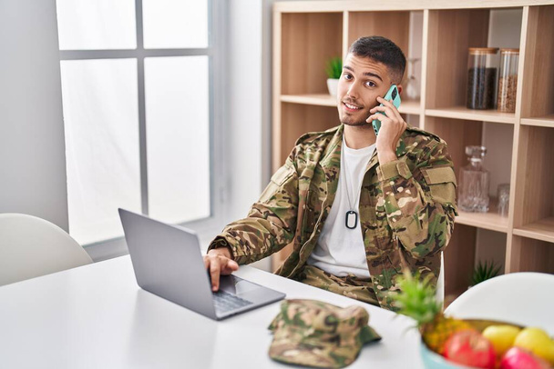 Young hispanic man army soldier using laptop talking on the smartphone at home - Photo, Image