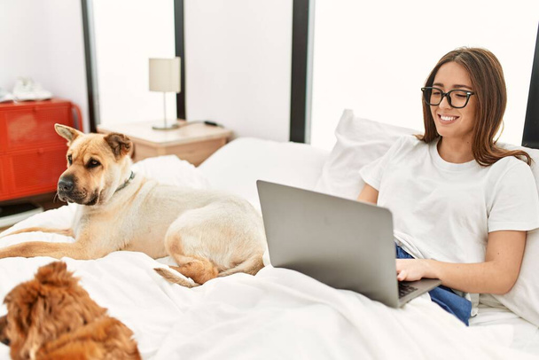 Mujer hispana joven usando portátil sentado en la cama con perros en el dormitorio - Foto, imagen