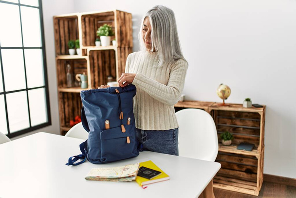Mujer de pelo gris de mediana edad sonriendo confiada preparando la mochila de viaje en casa - Foto, imagen