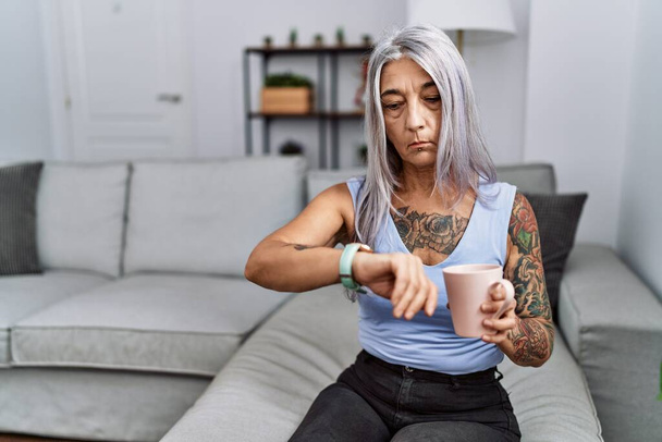 Mulher de cabelos grisalhos de meia idade bebendo café sentado no sofá em casa verificando a hora no relógio de pulso, relaxado e confiante  - Foto, Imagem