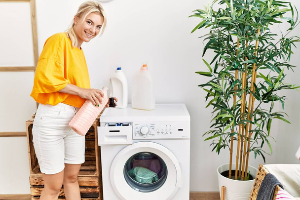 Joven chica rubia haciendo lavandería sosteniendo botella de detergente en casa. - Foto, Imagen