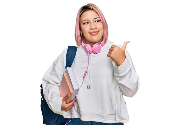 Mujer hispana con el pelo rosa usando mochila de estudiante y auriculares sonriendo con la cara feliz mirando y apuntando hacia un lado con el pulgar hacia arriba.  - Foto, imagen