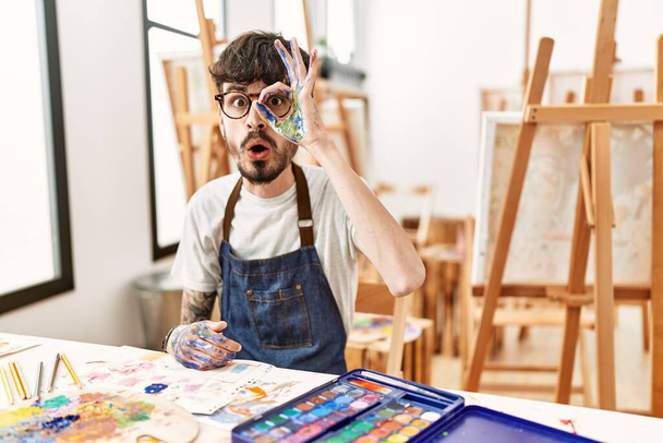 Homme hispanique avec barbe au studio d'art faisant un geste correct choqué par le visage surpris, les yeux regardant à travers les doigts. expression incrédule.  - Photo, image