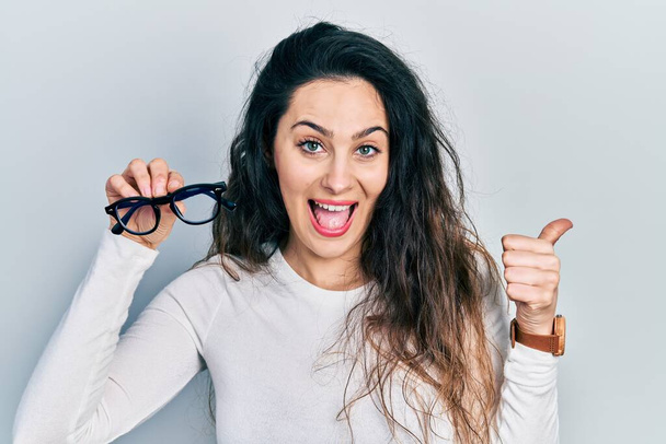 Молодая латиноамериканка в очках, указывая пальцем в сторону, улыбается счастливой с открытым ртом  - Фото, изображение