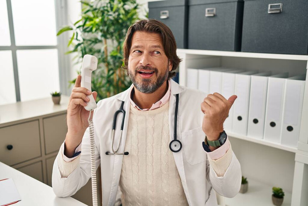 Handsome middle age doctor man speaking on the phone at the clinic pointing thumb up to the side smiling happy with open mouth  - 写真・画像