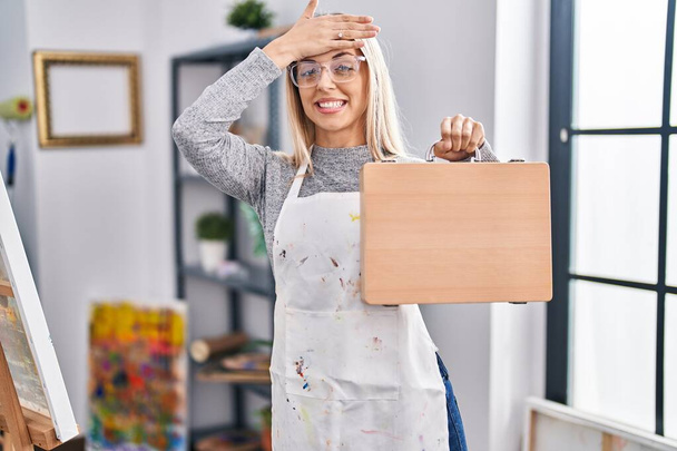 Mladá blondýna malíř žena drží dřevěné pouzdro v ateliéru stresu a frustrovaný s rukou na hlavě, překvapený a naštvaný obličej  - Fotografie, Obrázek