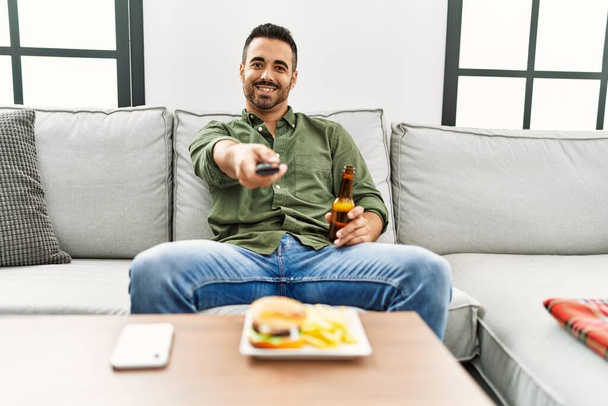 Young hispanic man eating hamburger watching tv at home - 写真・画像