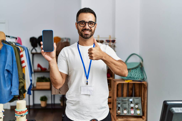 Junger hispanischer Mann mit Bart, der als Manager in einer Einzelhandelsboutique arbeitet und das Smartphone glücklich und positiv lächelnd in der Hand hält, Daumen hoch und Zustimmungszeichen  - Foto, Bild