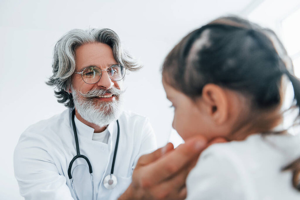 Little girl have a visit. Senior otorhinolaryngologist with grey hair and beard in white coat is indoors in clinic. - Foto, Imagem