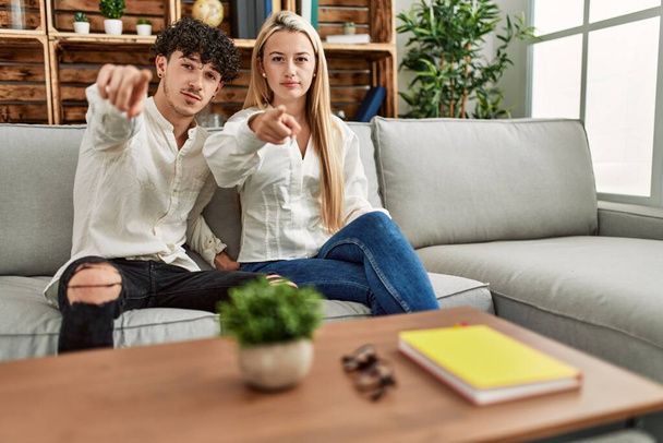 Young beautiful couple sitting on the sofa at home pointing with finger to the camera and to you, confident gesture looking serious  - 写真・画像