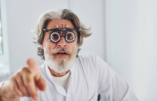 Nice facial expressions. Senior man with grey hair and beard is in ophthalmology clinic. - Photo, Image